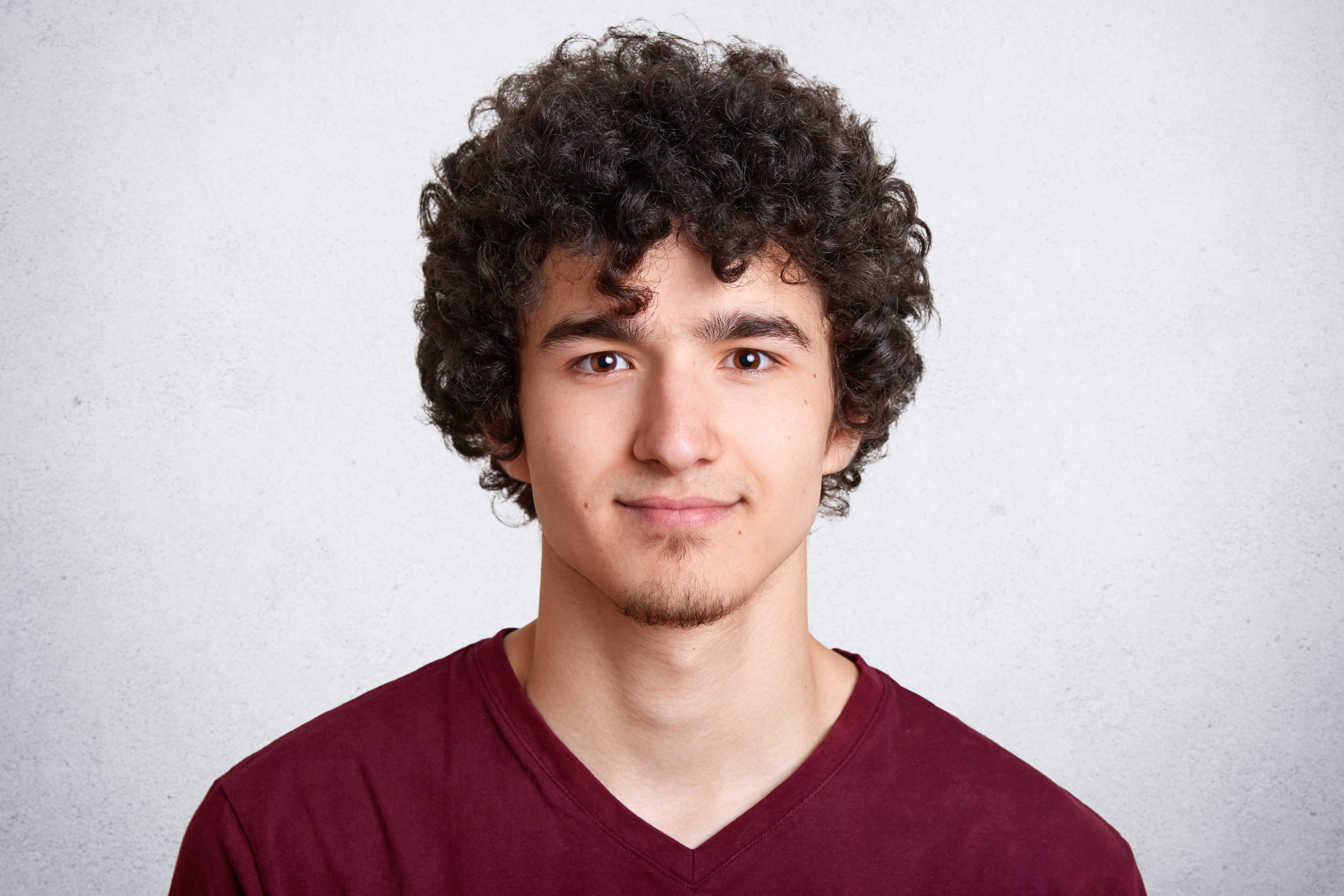 Close up portrait of curly handsome European male, wearing casual maroon shirt, isolated over white background, looking directly at camera, beig photographed in photo studio. People concept.